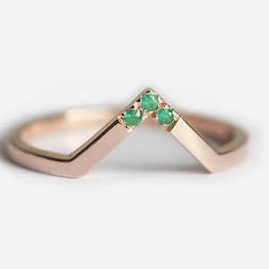 May include: A rose gold chevron ring with three small, round emeralds set at the point of the V. The ring is set against a white background, highlighting the polished metal and vibrant green gemstones.