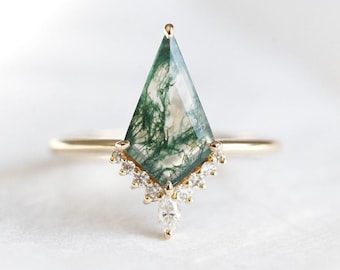 May include: A gold ring featuring a large, kite-shaped moss agate gemstone with green and white inclusions. The ring is accented with small, clear gemstones along the band and a single pear-shaped stone below the agate.