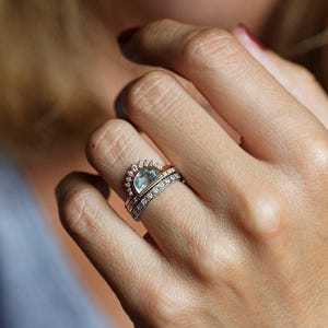 May include: A ring with a light blue gemstone surrounded by small diamonds. The ring is set in gold and has a delicate band. The ring is worn on a finger with a second, thinner band with diamonds.