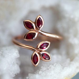 Puede incluir: Un anillo de oro rosa con dos ramas entrelazadas, cada una adornada con tres piedras preciosas de rubí rojo. El anillo está diseñado para ser usado en el dedo.