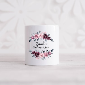 May include: White ceramic jar featuring a floral design in pink, burgundy, and grey. The text "Sarah's Sadaqah Jar" is displayed on the jar. The floral arrangement encircles the text, creating a decorative and elegant appearance.