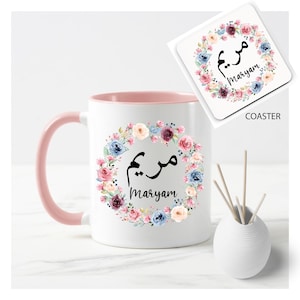 May include: A white mug with a pink rim and handle, featuring a floral wreath with the name "Maryam" written in Arabic calligraphy. The mug is accompanied by a matching coaster with the same design.