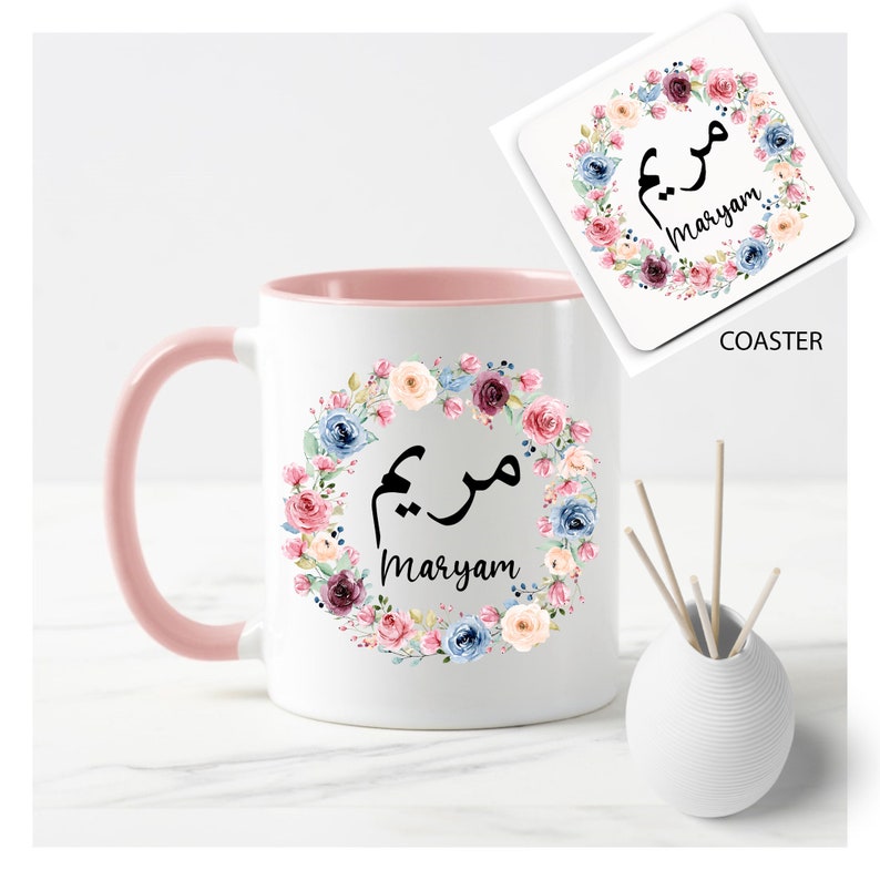 May include: A white mug with a pink rim and handle, featuring a floral wreath with the name "Maryam" written in Arabic calligraphy. The mug is accompanied by a matching coaster with the same design.