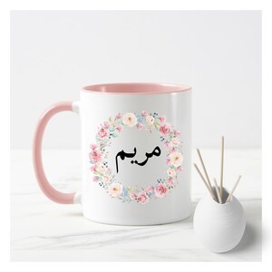 May include: A white ceramic mug with a pink rim and handle. The mug features a floral wreath design with pink, white, and blue flowers. The word "مريم" is written in black inside the wreath.