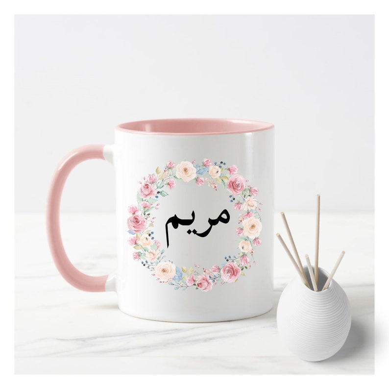May include: A white ceramic mug with a pink rim and handle. The mug features a floral wreath design with pink, white, and blue flowers. The word "مريم" is written in black inside the wreath.