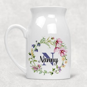 May include: White ceramic mug featuring a floral wreath design. The wreath is composed of pink, purple, and yellow flowers and green leaves. The letter "N" is in the centre of the wreath, with the word "Nanny" written below it.