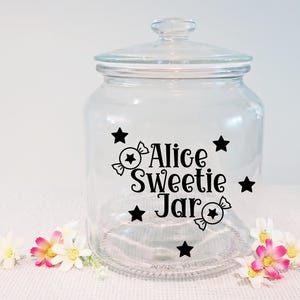 May include: A clear glass jar with a lid, decorated with black stars and candy graphics. The text "Alice Sweetie Jar" is displayed on the front. The jar is sitting on a white surface with flowers.