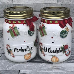 May include: Two white mason jars decorated with red and white Christmas designs. The jars are labelled "Marshmallows" and "Hot Chocolate".