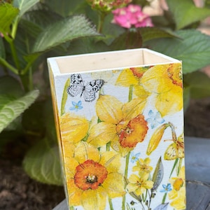 May include: A white wooden box with a floral design featuring yellow daffodils and a black and white butterfly. The box has a light wood top and a white background with a floral pattern.