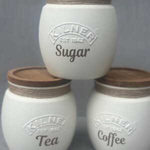 May include: Three off-white ceramic storage jars with wooden lids and rope detailing. The jars are labelled "Sugar", "Tea", and "Coffee" in brown lettering. Each jar has the "Kilner Est 1842" logo.