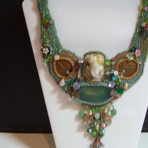 May include: A beaded necklace with a green, brown, and gold color scheme. The necklace features a large, light green stone pendant and a smaller, white stone pendant. The necklace is decorated with various beads, including pearls, crystals, and flowers.