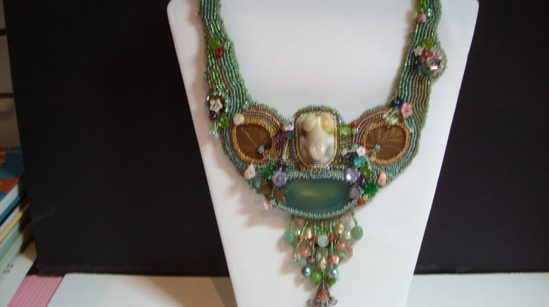 May include: A beaded necklace with a green, brown, and gold color scheme. The necklace features a large, light green stone pendant and a smaller, white stone pendant. The necklace is decorated with various beads, including pearls, crystals, and flowers.