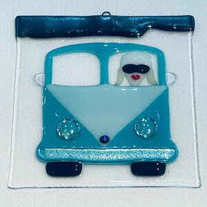 May include: A turquoise and light blue glass art piece featuring a stylized van with a driver wearing sunglasses and a red mouth. The van has clear glass accents and black tires. A dark blue border and a loop for hanging complete the design.