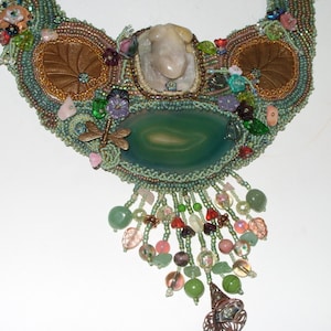 May include: A green and brown beaded necklace with a large green agate stone pendant and a smaller white stone. The necklace has a fringe of beads and is decorated with small flowers and other embellishments.