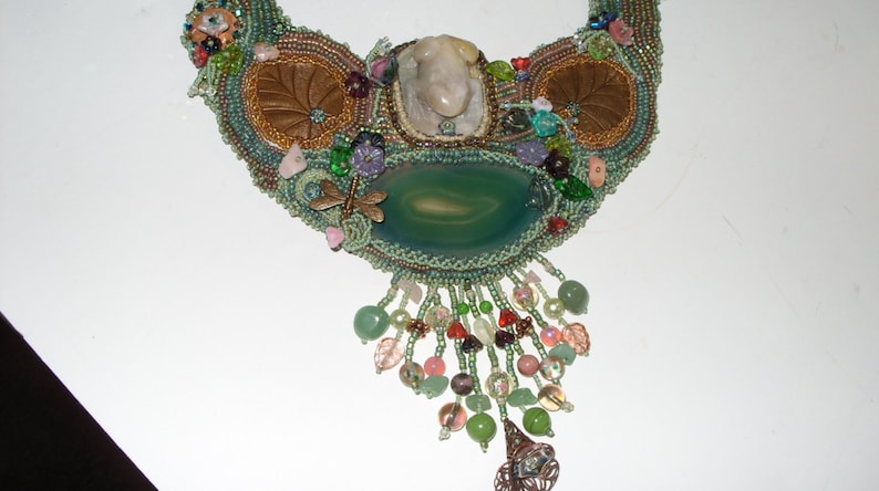 May include: A green and brown beaded necklace with a large green agate stone pendant and a smaller white stone. The necklace has a fringe of beads and is decorated with small flowers and other embellishments.