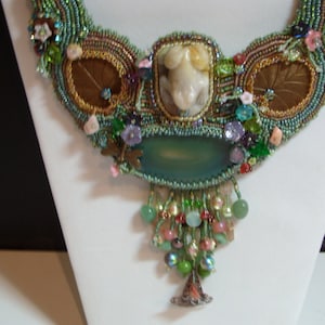 May include: A beaded necklace with a green and brown color scheme. The necklace features a large, light green stone, a smaller white stone, and several smaller beads in various colors. The necklace is displayed on a white stand.