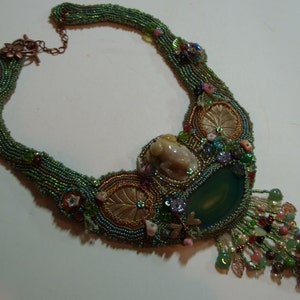 May include: A green, blue, and gold beaded necklace with a large green agate stone pendant. The necklace features a small, white ceramic frog charm and intricate beadwork.