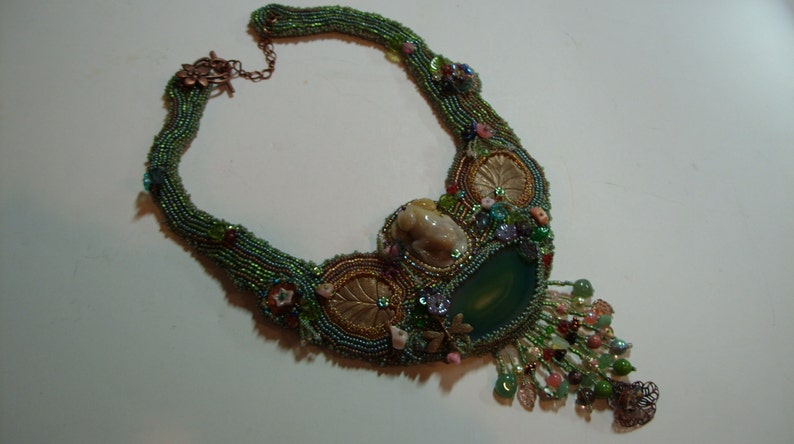 May include: A green, blue, and gold beaded necklace with a large green agate stone pendant. The necklace features a small, white ceramic frog charm and intricate beadwork.
