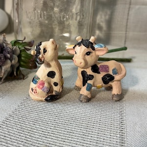 May include: Two small, hand-painted cow figurines. One is sitting, the other standing. Both have black spots and patches of color. The figurines are on a striped surface, with dried lavender and a vase in the background.