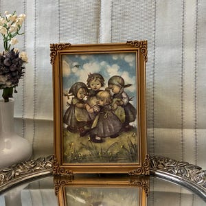 May include: A vintage framed print featuring a group of children in a field, with a gold-colored ornate frame. The image is set on a mirrored tray, with a vase of flowers and a textured vase in the background.