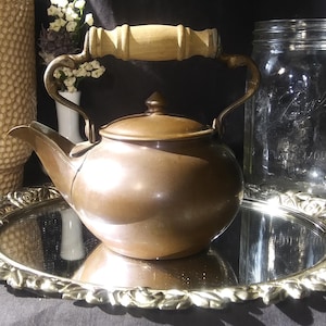 May include: A copper teapot with a wooden handle sits on a gold-colored tray with a mirrored surface. The teapot has a rounded body and a spout on the side. There is a glass jar and a ceramic vase in the background.