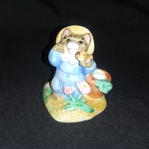 May include: A ceramic figurine of a cat wearing a straw hat and a blue dress, holding a basket of flowers. The cat is standing on a grassy base.
