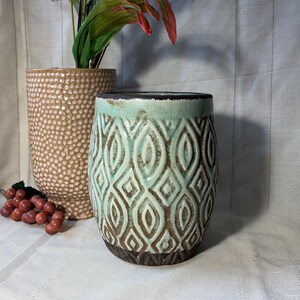 May include: A ceramic vase with a teal and brown geometric pattern. The vase has a cylindrical shape with a dark brown base and rim. A second vase with a textured surface is behind it. Decorative items.