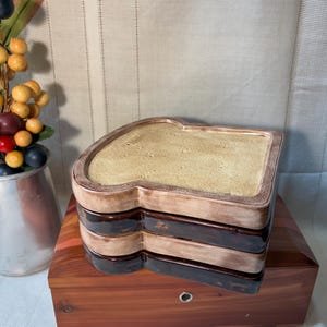 May include: A stack of three ceramic plates shaped like slices of bread, featuring a textured tan center and a dark brown crust edge. The plates are layered on top of a rectangular wooden box, with a metal pitcher of artificial berries nearby.