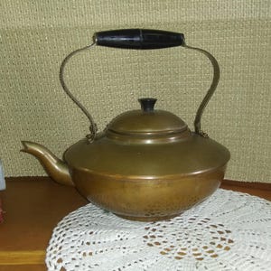 May include: A vintage copper tea kettle with a black handle and lid. The kettle has a spout and a curved handle. It sits on a white lace doily. The kettle is a warm, antique color.