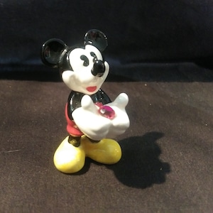 May include: A small, ceramic Mickey Mouse figurine. Mickey is depicted in his classic attire: black ears and body, white gloves, red shorts, and yellow shoes. He holds a pink gem in his hands.