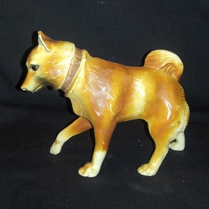 May include: A brown ceramic figurine of a dog with a brown leather collar. The dog is standing on all four paws and has a long tail.