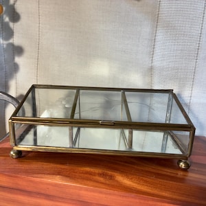 May include: A rectangular glass display box with a brass metal frame and a mirrored base. The box is divided into three separate compartments by two internal glass dividers. The lid features a delicate etched floral design and rests on four small ball feet.
