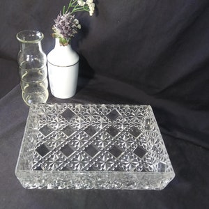 May include: A clear glass rectangular dish with a diamond pattern design. The dish is sitting on a black surface. There are two clear glass vases and a white ceramic vase with a gold trim in the background.