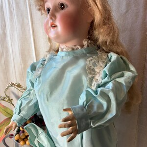 Op de afbeelding: Een vintage porseleinen pop met blond haar en roze wangen. De pop draagt een lichtblauwe satijnen jurk met kanten afwerking. De armen van de pop zijn uitgestrekt en ze heeft een serene uitdrukking.