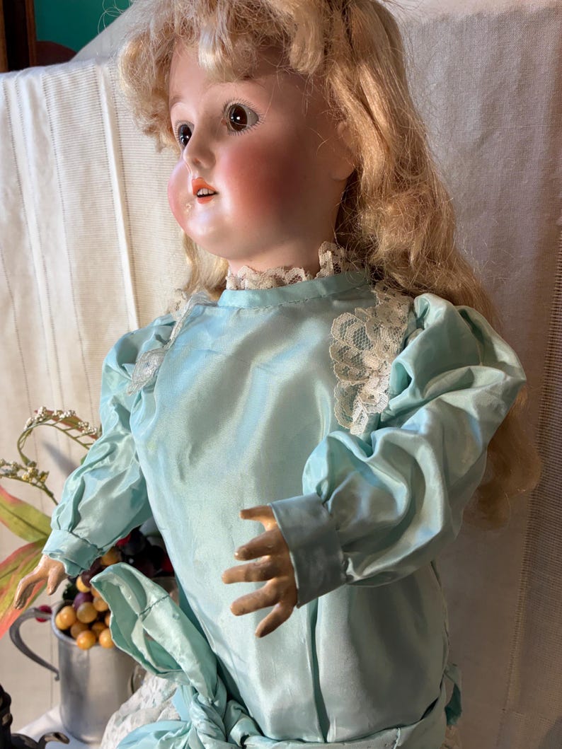 Op de afbeelding: Een vintage porseleinen pop met blond haar en roze wangen. De pop draagt een lichtblauwe satijnen jurk met kanten afwerking. De armen van de pop zijn uitgestrekt en ze heeft een serene uitdrukking.