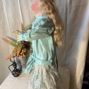 Op de afbeelding: Een vintage pop met lang blond haar, gekleed in een lichtblauwe satijnen jurk met een witte kanten rok. De pop houdt bloemen vast. Op de tafel staat een kleine, decoratieve zwarte theepot.