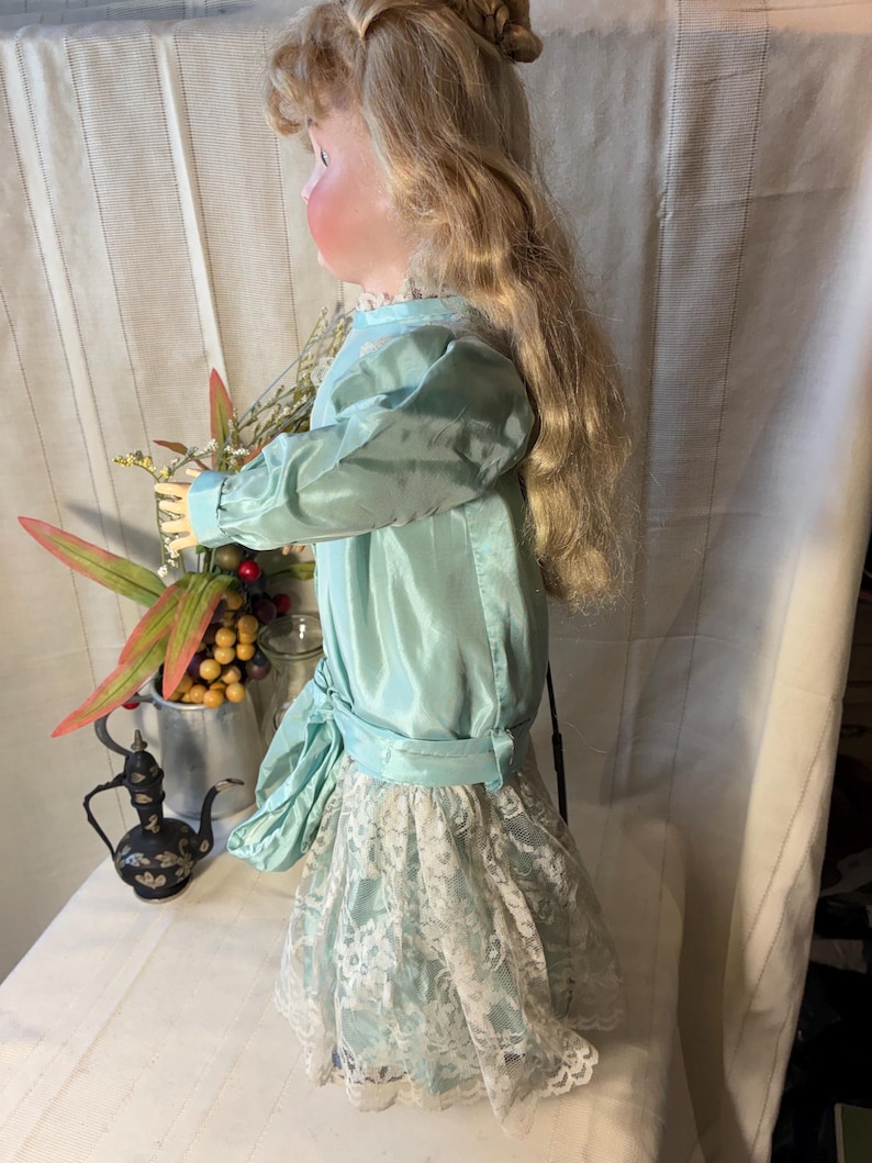 Op de afbeelding: Een vintage pop met lang blond haar, gekleed in een lichtblauwe satijnen jurk met een witte kanten rok. De pop houdt bloemen vast. Op de tafel staat een kleine, decoratieve zwarte theepot.