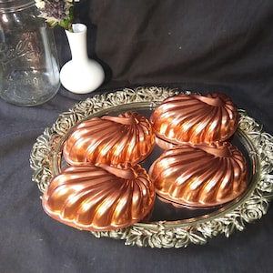 May include: Four copper-colored, fluted molds sit on a silver-colored, ornate tray. A clear glass jar and a small white vase with flowers are in the background. The molds have a shiny, reflective surface.