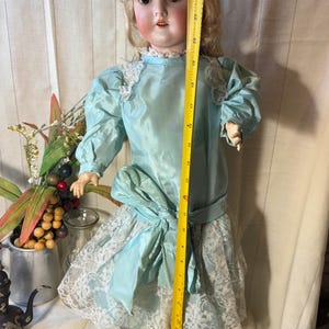 Op de afbeelding: Antieke porseleinen pop in een lichtblauwe jurk met kanten afwerking. De pop is ongeveer 58 cm hoog. De jurk heeft een grote strik in de taille en pofmouwen. De pop heeft blond haar en een geschilderd gezicht.