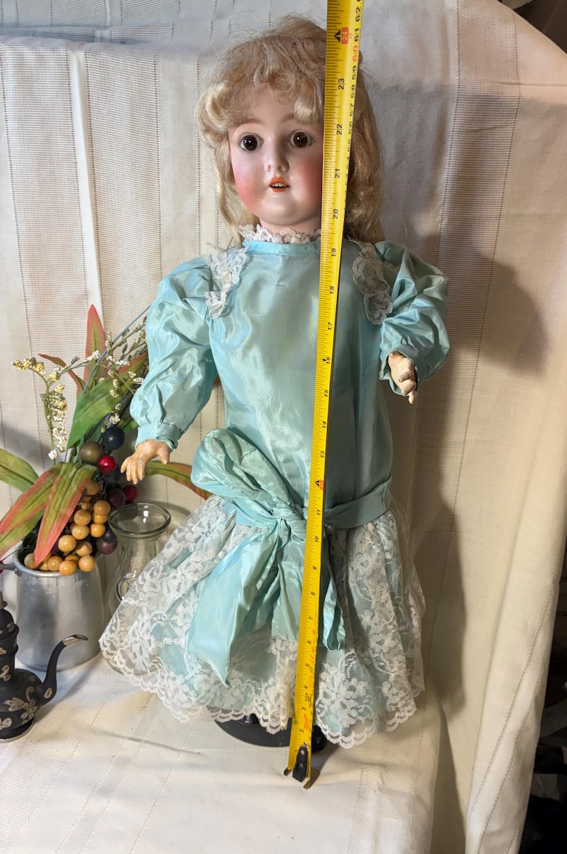 Op de afbeelding: Antieke porseleinen pop in een lichtblauwe jurk met kanten afwerking. De pop is ongeveer 58 cm hoog. De jurk heeft een grote strik in de taille en pofmouwen. De pop heeft blond haar en een geschilderd gezicht.