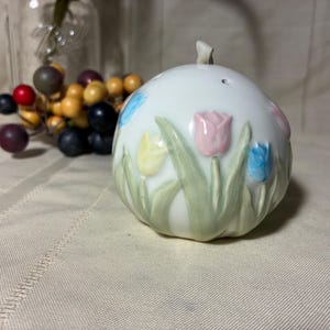 May include: A white ceramic salt shaker with a floral design featuring pink, blue, and yellow tulips. The shaker has a small handle on top and is decorated with green leaves. The background includes a glass jar and decorative berries.