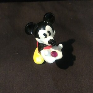 May include: A figurine of Mickey Mouse, a classic Disney character, in a seated pose. Mickey is depicted with black ears, a white face, and a red bow tie. He holds a white hand with a pink gem. His shoes are yellow.
