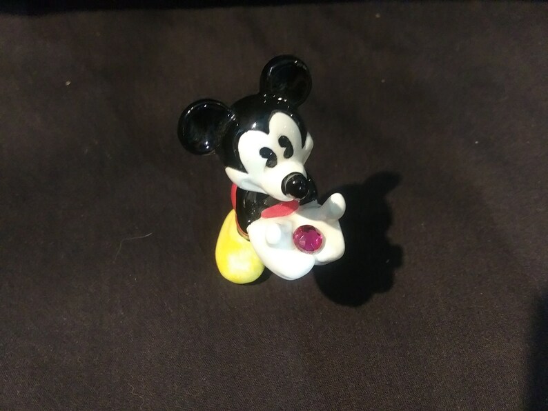 May include: A figurine of Mickey Mouse, a classic Disney character, in a seated pose. Mickey is depicted with black ears, a white face, and a red bow tie. He holds a white hand with a pink gem. His shoes are yellow.