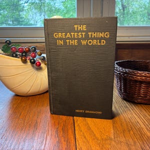 May include: A black hardcover book with gold lettering, titled "THE GREATEST THING IN THE WORLD" by Henry Drummond. It stands on a wooden surface. A small woven basket and a decorative bowl with berries are also visible.