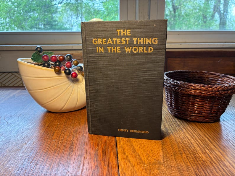 May include: A black hardcover book with gold lettering, titled "THE GREATEST THING IN THE WORLD" by Henry Drummond. It stands on a wooden surface. A small woven basket and a decorative bowl with berries are also visible.