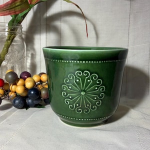 May include: A dark green ceramic planter with a decorative floral design. The planter has a slightly flared shape and a textured surface. The design is centered on the front of the planter and is surrounded by a dotted border.