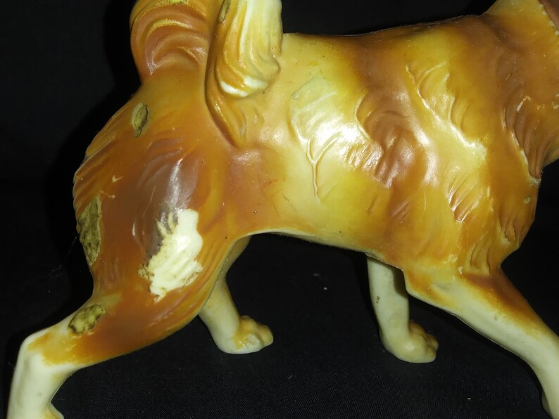 May include: A ceramic figurine of a brown and white dog with a long tail. The dog is standing on all four legs and has a slightly tilted head.