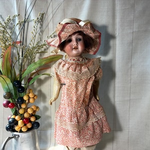 May include: A vintage doll with brown hair wearing a matching peach and white floral patterned dress and bonnet with lace trim. The doll has painted facial features and is positioned next to a silver pitcher filled with decorative berries and foliage.
