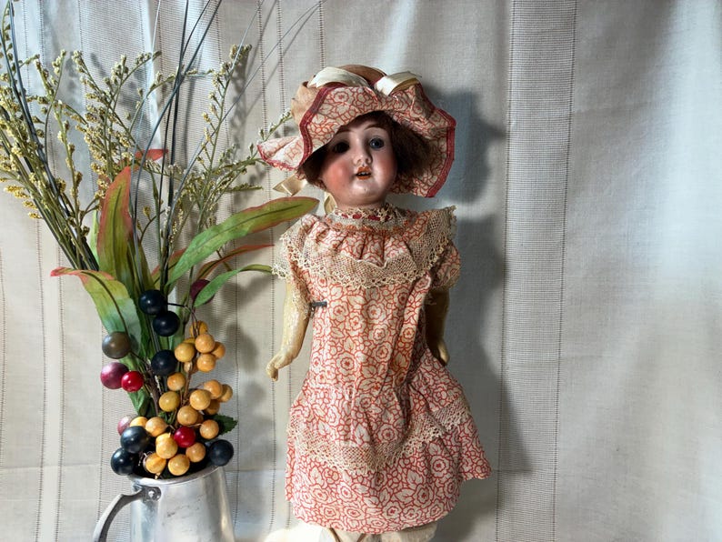 May include: A vintage doll with brown hair wearing a matching peach and white floral patterned dress and bonnet with lace trim. The doll has painted facial features and is positioned next to a silver pitcher filled with decorative berries and foliage.