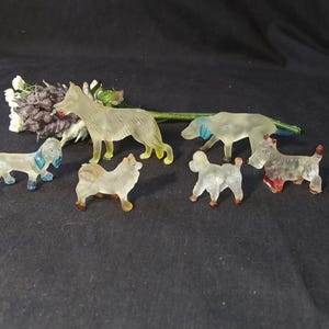 May include: A collection of six clear plastic dog figurines. The dogs are in various poses and sizes, and some have painted details in blue, red, and yellow.